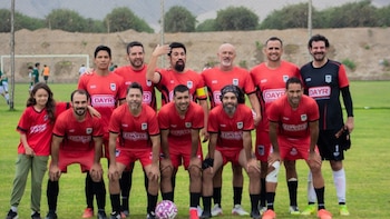 Grupo de once hombres y una niña posan sonrientes en un campo de fútbol. La mayoría lleva uniformes rojos, uno de portero negro y rojo