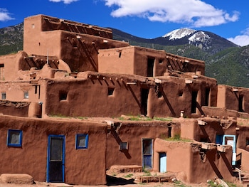 Conjunto de edificaciones de color ocre construidas con adobe, con puertas y ventanas. Fondo de montañas con vegetación, un pico nevado y cielo azul con nubes