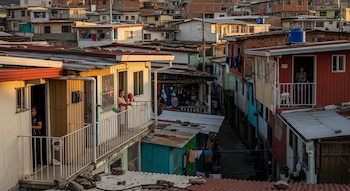 Vista aérea de un barrio denso con casas de colores variados y techos de metal. Varias personas se asoman desde balcones y puertas.