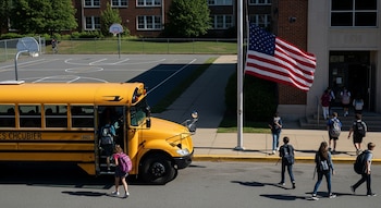 Alumnos llegan a un colegio en Estados Unidos, con un autobús escolar amarillo, una bandera estadounidense y una cancha de baloncesto visible en el campus.
