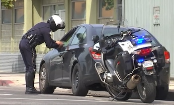 Un agente de policía con casco blanco habla por la ventanilla de un auto gris junto a una motocicleta policial