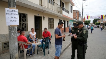 Dos policías imponen un comparendo a un hombre con una botella de cerveza en una calle de Colombia, con un cartel de "Ley Seca Vigente" y personas bebiendo.