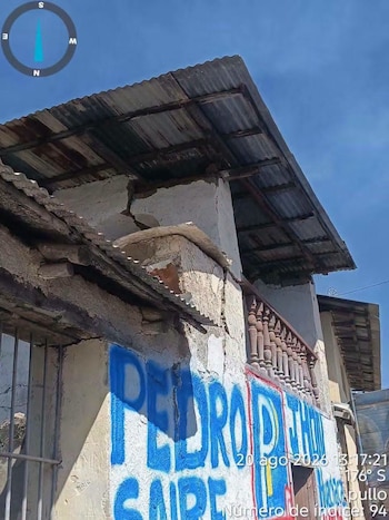 Fachada de una vivienda con grietas, derrumbe parcial y graffiti azul, un techo de chapa, balcón de madera y una brújula en la esquina superior