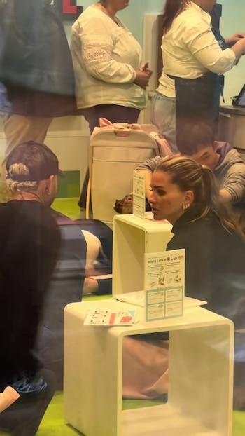Eugenia 'La China' Suárez, con cabello recogido y vestida de negro, sentada en un café de minipigs en Japón, con otras personas y señalización