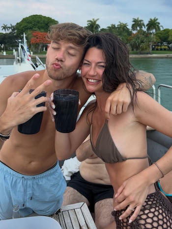 Un hombre rubio con barba y una mujer de cabello oscuro posan a bordo de un yate, sostienen vasos, con palmeras y agua de fondo