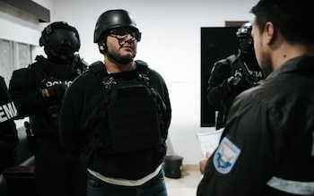 Un hombre con casco negro, gafas, sudadera oscura y chaleco táctico está de pie, custodiado por tres agentes de policía con uniformes y equipo táctico