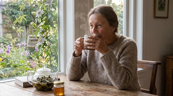 Mujer con suéter claro bebe de una taza humeante. Una tetera de vidrio con pasiflora y un tarro de miel en mesa de madera junto a una ventana con jardín.