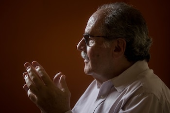 Un hombre con gafas, bigote y camisa de rayas gesticula con las manos levantadas ante un fondo de tono oscuro