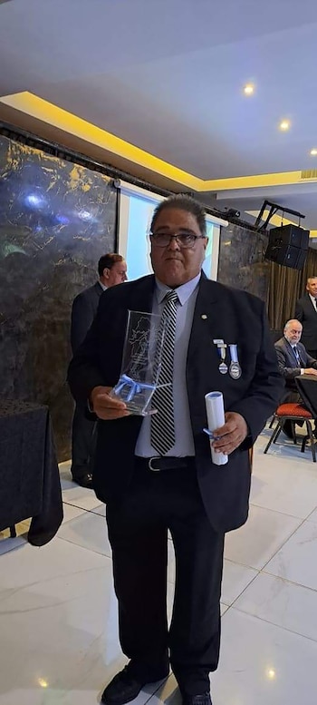 Hombre de mediana edad con traje oscuro, corbata y gafas, sostiene un premio transparente y un certificado blanco enrollado. Otros asistentes en el fondo