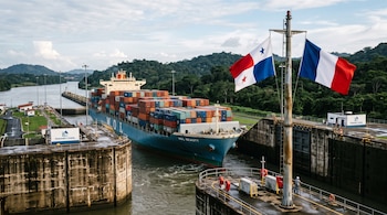 Un buque de carga azul lleno de contenedores cruza una esclusa de concreto. A la derecha, dos banderas, de Panamá y Francia, flamean en un mástil.