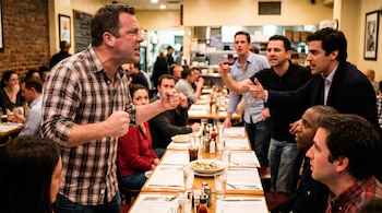 Un hombre de piel normal con camisa a cuadros grita con los puños cerrados en un restaurante. Otros hombres discuten y señalan mientras los comensales observan.