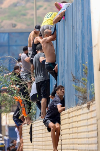 Varios hombres escalan una valla de metal corrugado azul y una pared beige. Algunos hombres están en la parte superior, otros en la parte inferior o media