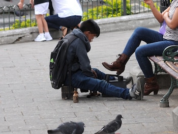 Un niño con mochila y jeans lustra los zapatos de una mujer sentada en un banco de parque. En el suelo hay palomas y una botella pequeña