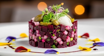 Un plato blanco contiene un tartar cilíndrico de remolacha y rábanos cortados en dados, coronado con alcaparras, pepinillos, brotes y crema, decorado con pétalos de flores y salsa.