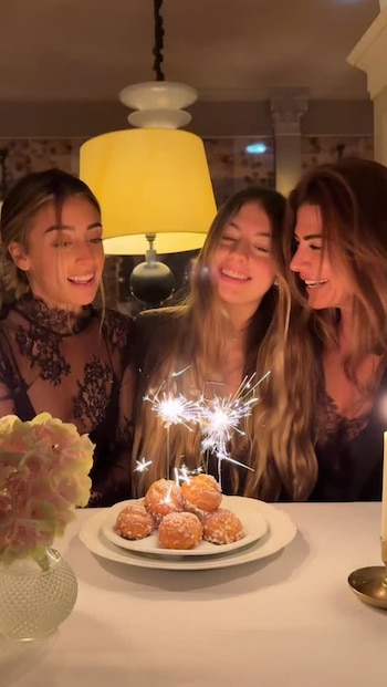 Tres mujeres sonríen frente a un plato de buñuelos con bengalas encendidas sobre una mesa