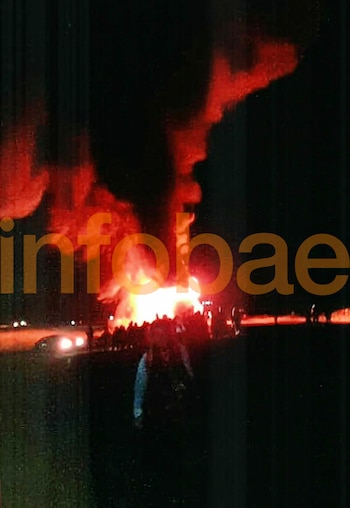 Incendio de vehículo en carretera con grandes llamas y humo denso en la noche. Siluetas de personas observan. Luces de automóviles presentes. Logotipo de Infobae