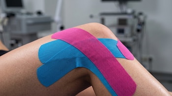 Tiras de cinta elástica azul y rosa adheridas sobre la rodilla flexionada de una persona, con equipo médico desenfocado en el fondo.