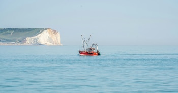 Un barco pesquero de color rojo navega en un mar calmo frente a acantilados blancos bajo un cielo claro