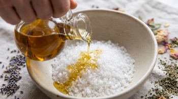 Una mano sujeta una jarra de vidrio y vierte aceite de oliva dorado sobre un montículo de sal gruesa en un cuenco de cerámica, con flores secas y hierbas.