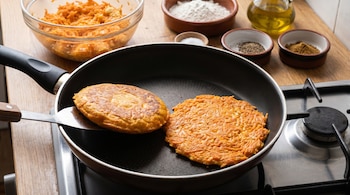 Dos tortillas de zanahoria cocinándose en una sartén antiadherente sobre una estufa. Una está dorada y la otra, anaranjada. En el fondo, un bol con zanahoria rallada y huevos, y cuencos con harina, aceite, sal, pimienta y comino.