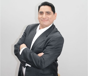 Un hombre de mediana edad, Carlos Álvarez, sonríe a la cámara con los brazos cruzados, vistiendo un traje a rayas oscuras y camisa blanca