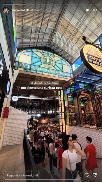 Ivana Icardi, de espaldas y con gafas de sol, camina por un concurrido mercado interior con techo alto de metal y un gran ventanal de vitral