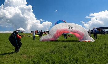 Campo verde con personas, varias velas de parapente extendidas, una carpa blanca y un cielo azul con nubes grandes