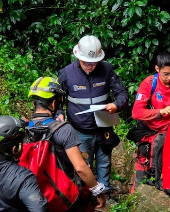 Tres personas con equipo de rescate: dos con cascos, una con chaleco de Protección Civil leyendo documentos, entre vegetación densa