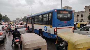 Autobús azul y blanco detenido en el tráfico, con luces de freno rojas, entre automóviles, motocicletas y auto rickshaws en calle urbana con cielo nublado.