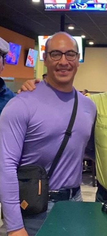 Juan Díaz, hombre con gafas y camiseta morada de manga larga, sonríe a la cámara con un bolso bandolera negro sobre su hombro en un interior