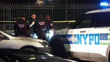 Tres agentes del NYPD con uniformes oscuros de pie cerca de un coche blanco y un SUV policial blanco y azul con luces azules encendidas, de noche