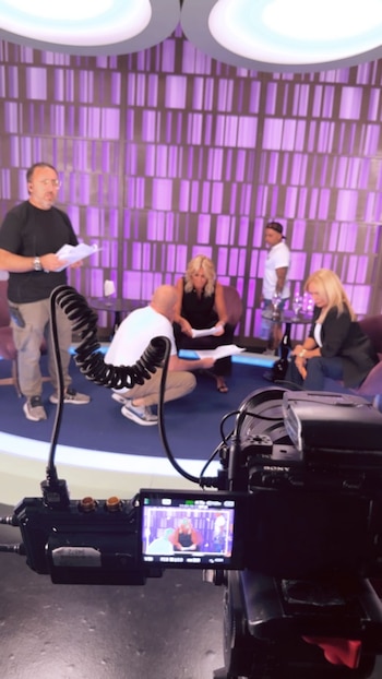 Vista detrás de escena de un set de grabación con cámara en primer plano. Cuatro personas, incluyendo Wanda Nara, Yanina Latorre y Pía Shaw, interactúan con guiones en un estudio violeta