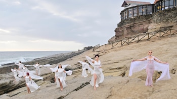 Diez mujeres con vestidos blancos posan sobre rocas, con el mar a la izquierda y un edificio de piedra en un acantilado a la derecha