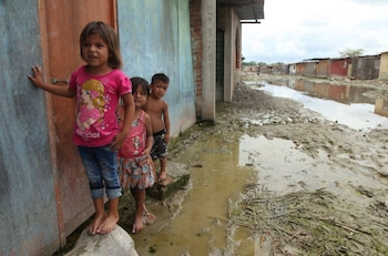 Tres niños descalzos parados sobre piedras en una calle inundada y embarrada junto a casas de madera y ladrillo
