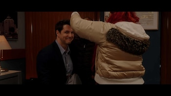 Un hombre sonriente con traje azul mira hacia la izquierda. A su derecha, una persona con cabello rojo y chaqueta acolchada crema con cuello de piel está de espaldas