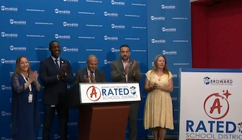 Cinco personas aplauden frente a un podio con el texto 'Broward County Public Schools Rated A School District'. El fondo es azul con logotipos