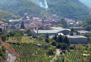 Panorámica de un pueblo italiano en un valle verde, con una iglesia de cúpulas, casas, edificios industriales, y humo ascendiendo en la ladera