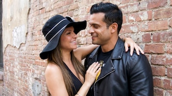 Un hombre y una mujer sonrientes se miran, con la mujer usando un sombrero negro y el hombre una chaqueta de cuero, frente a una pared de ladrillo.