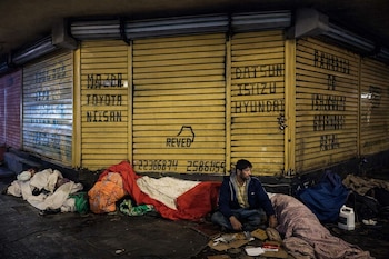 Un hombre se sienta en el suelo junto a mantas y cartones, frente a persianas metálicas amarillas con grafitis que incluyen nombres de marcas de vehículos.
