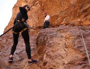 Mujer con casco y arnés de rapel en primer plano. Dos hombres con casco al fondo. Cuerdas de escalada. Pared rocosa de color rojizo
