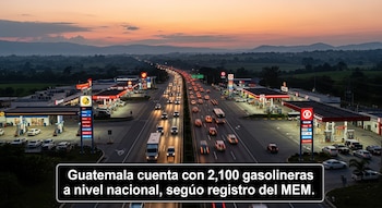 Vista aérea de una autopista con tráfico de vehículos y luces encendidas al atardecer, flanqueada por gasolineras iluminadas y montañas al fondo.