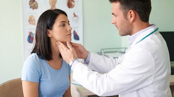 Un médico con bata blanca examina con las manos el cuello de una mujer joven con camiseta azul en un consultorio. Un diagrama anatómico se ve al fondo.
