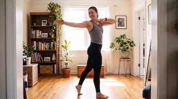 Mujer descalza con top gris y mallas negras estira los brazos en un salón con estantería de libros, plantas, una ventana grande y suelo de madera.