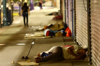 Varias personas, incluyendo una mujer, duermen en la acera sobre cartones junto a persianas metálicas de comercios cerrados en una calle oscura por la noche
