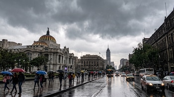 Personas con paraguas caminan por una acera mojada junto al Palacio de Bellas Artes y una avenida con vehículos bajo un cielo gris.