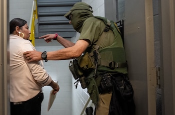 Un agente de United States Immigration and Customs Enforcement (ICE) con equipo táctico verde y rostro cubierto señala a una mujer en un pasillo interior