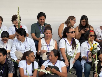 Un grupo de personas sentadas en gradas viste prendas blancas y cintas moradas mientras sostiene flores y teléfonos móviles