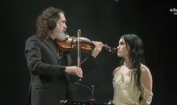 Lali Espósito, con cabello oscuro y vestida de claro, mira a un hombre barbudo con auriculares tocando el violín en un escenario oscuro
