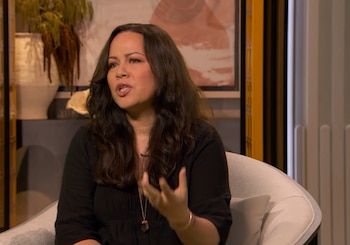 Mujer con cabello oscuro y blusa negra sentada en un sillón, gesticula con la mano derecha. Usa un collar y un brazalete. Fondo con plantas y cuadros