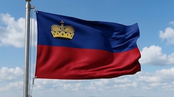 Bandera de Liechtenstein (azul y roja con una corona dorada) ondeando en un mástil gris. Cielo azul con nubes, montañas y un pueblo se ven al fondo